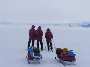 Tre skiløpere med pulk ser mot fjell i Antarktis.